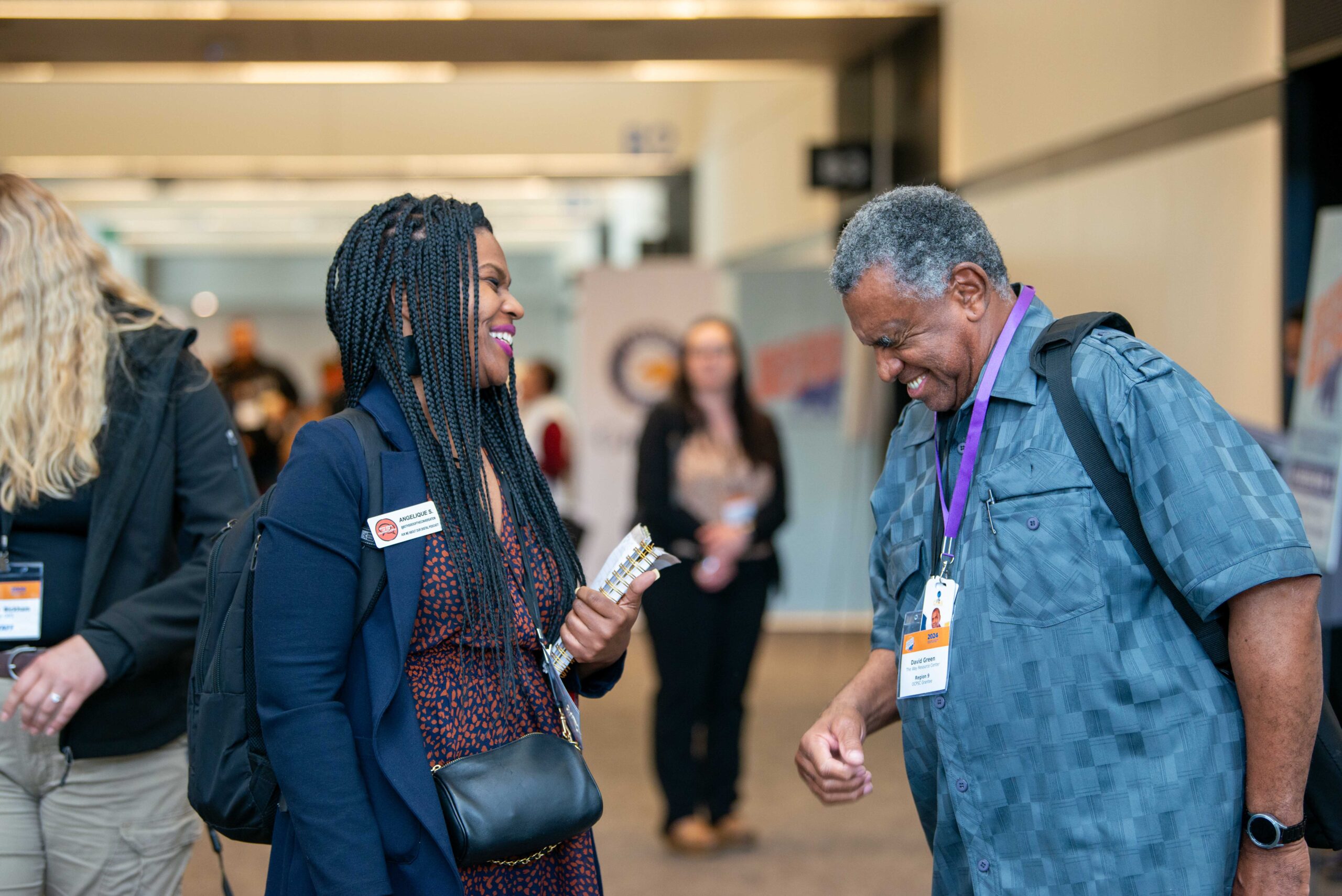 Two conference attendees wearing name badges smile and talking in a hallway while others network in the background, representing connection and engagement.
