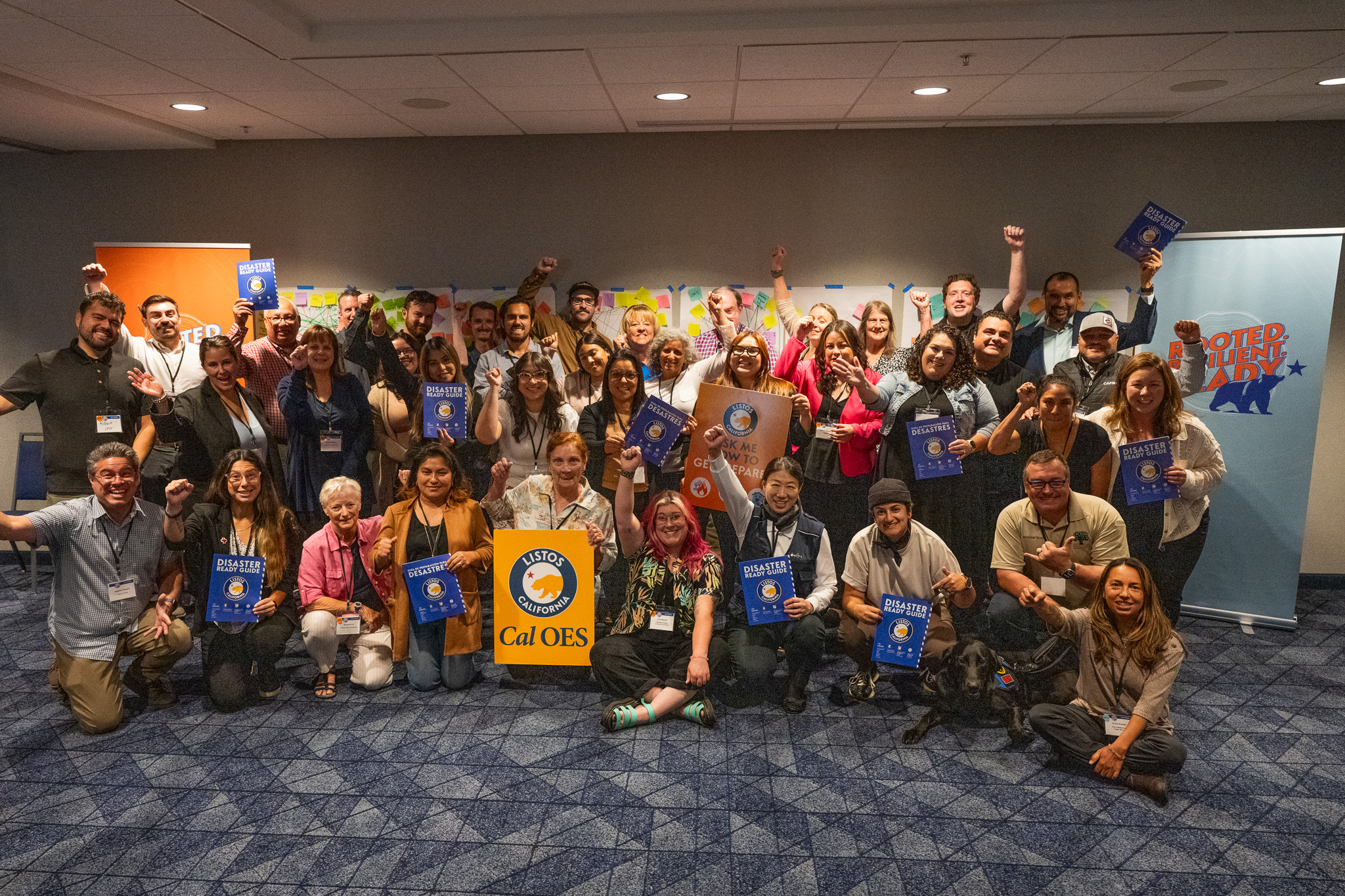 A large, diverse group people pose together indoors during a Listos Summit, smiling and celebrating while holding Listos signs. 