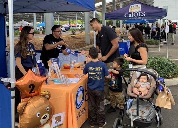 Cal OES Listos staff engage with community members and families at an outdoor event, standing at an informational table with outreach materials.