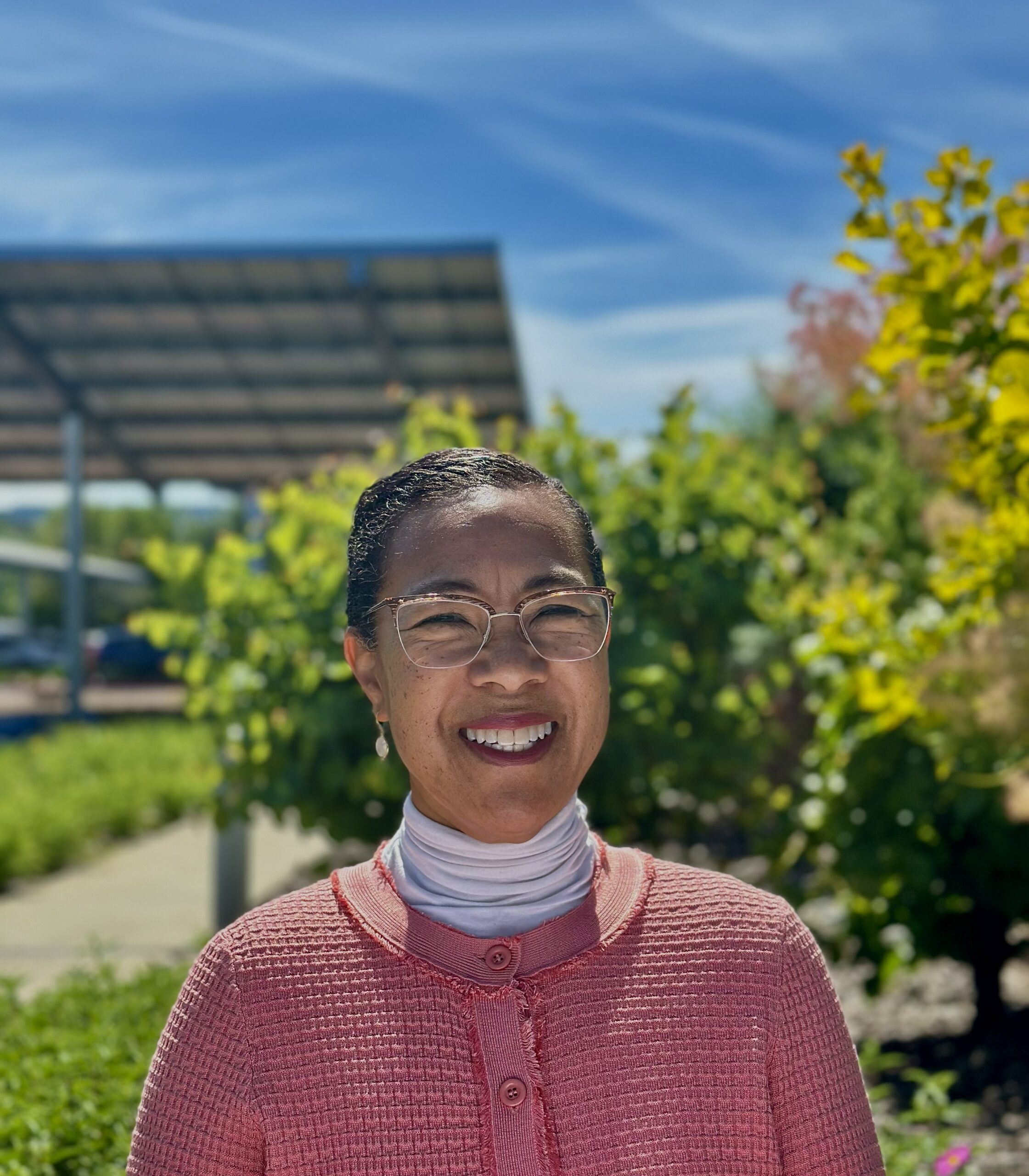 Portrait of Monisha Avery, Chief Equity Officer at Cal OES. Monisha is smiling and wearing glasses. The picture was taken outdoors in a sunny garden setting with green plants in the background under a bright blue sky.