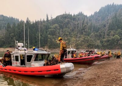 Firefighters getting onto three fire boats from the shore of a lake.